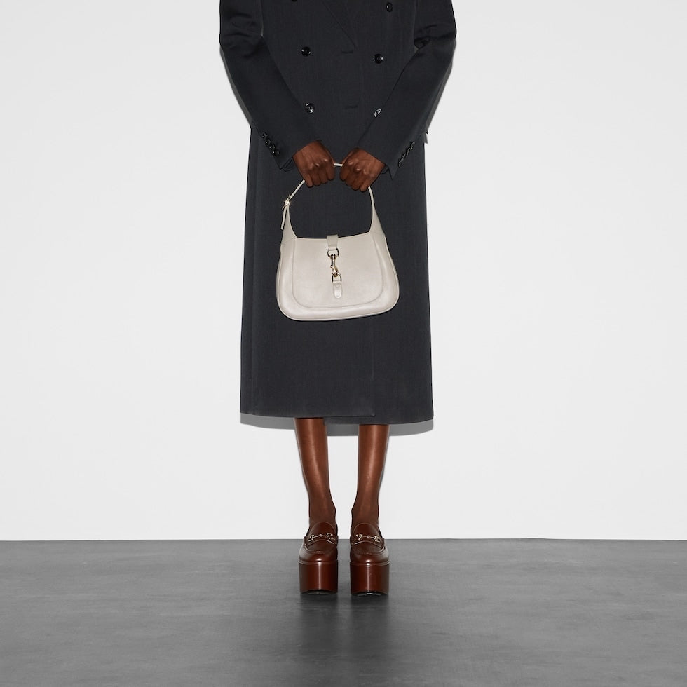 Person holding a white handbag wearing a long dark coat and brown shoes on a plain background