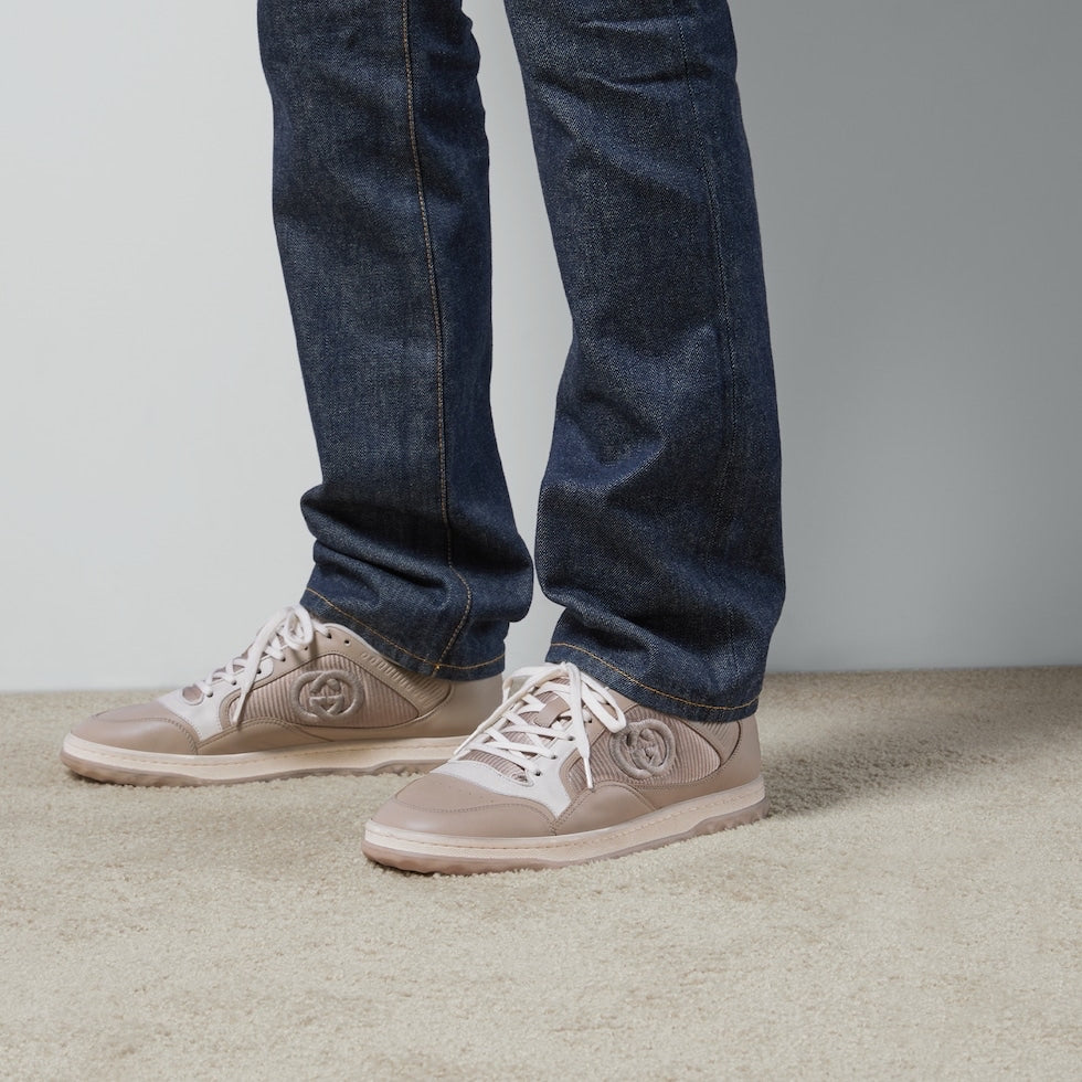 Person wearing blue jeans and beige sneakers with a Gucci logo on a neutral background