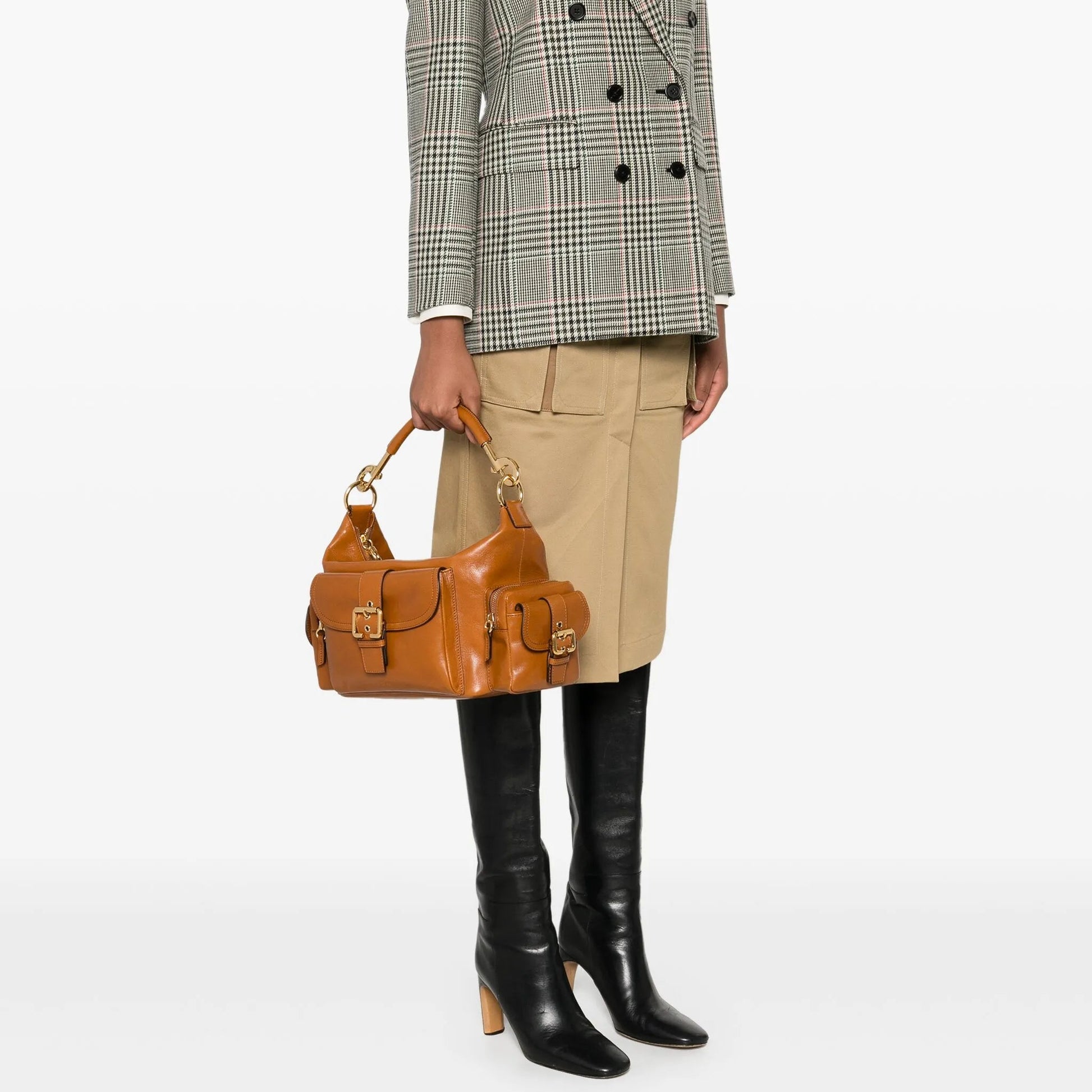 Person wearing a plaid blazer, beige skirt, black boots, and holding a brown handbag on a white background