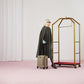 Person in a gray cape holding a suitcase next to a gold luggage stand on a pink floor.