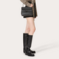 Person wearing a patterned shirt, black skirt, and black knee-high boots holding a black handbag on a white background