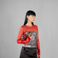 Woman wearing a red long-sleeve top with a silver sequin top underneath, holding a patterned handbag against a gray background.