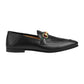 Black loafer with a gold and green horsebit detail on a light gray background