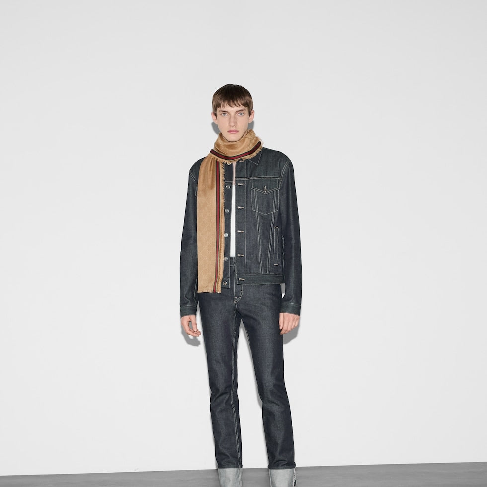 Person wearing a dark denim jacket and jeans with a beige scarf on a white background