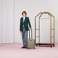 Man in a green suit holding a suitcase next to a gold luggage rack on a pink floor.