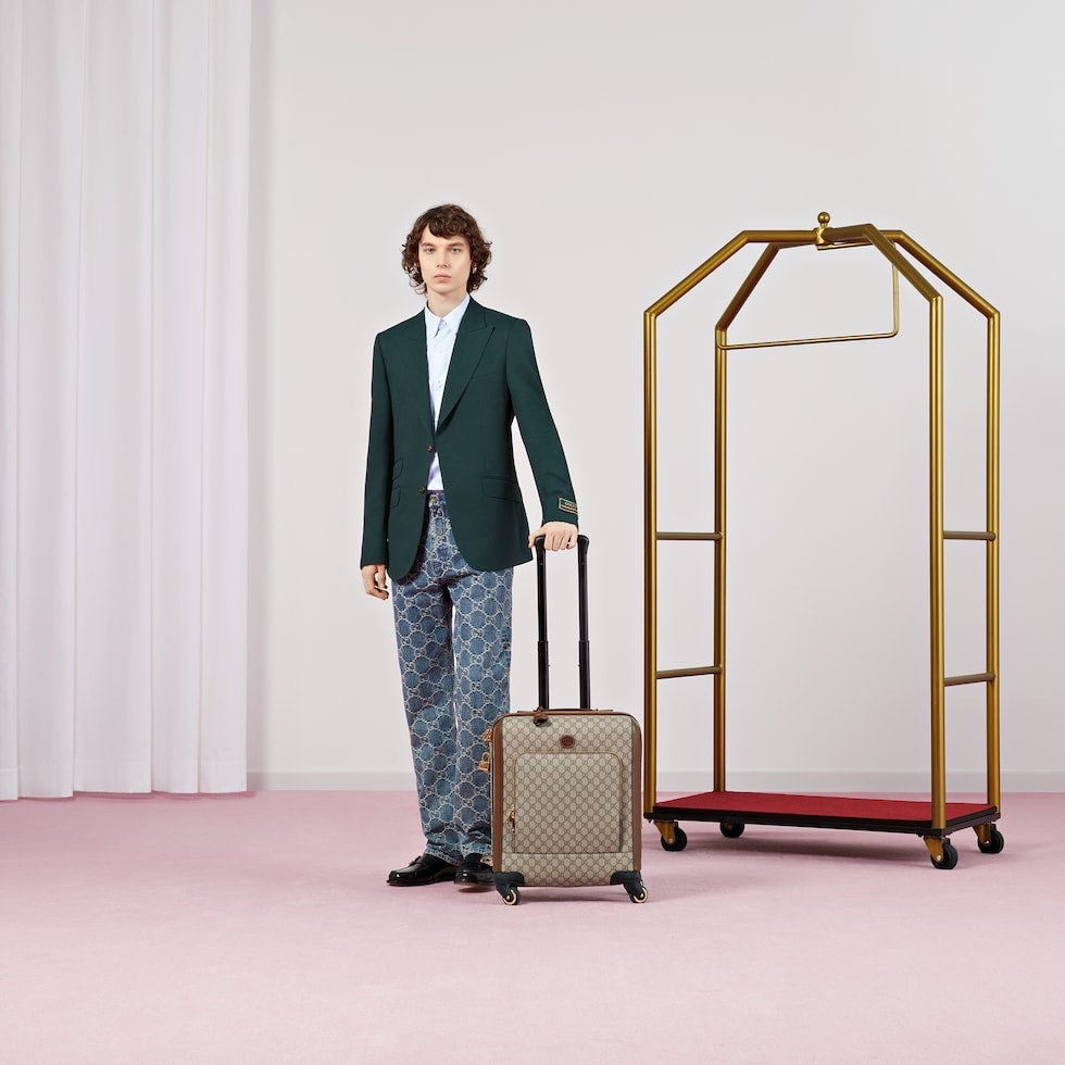 Man in a green suit holding a suitcase next to a gold luggage rack on a pink floor.