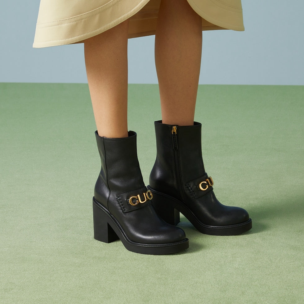 Black ankle boots with gold buckles worn by a person on a green floor.
