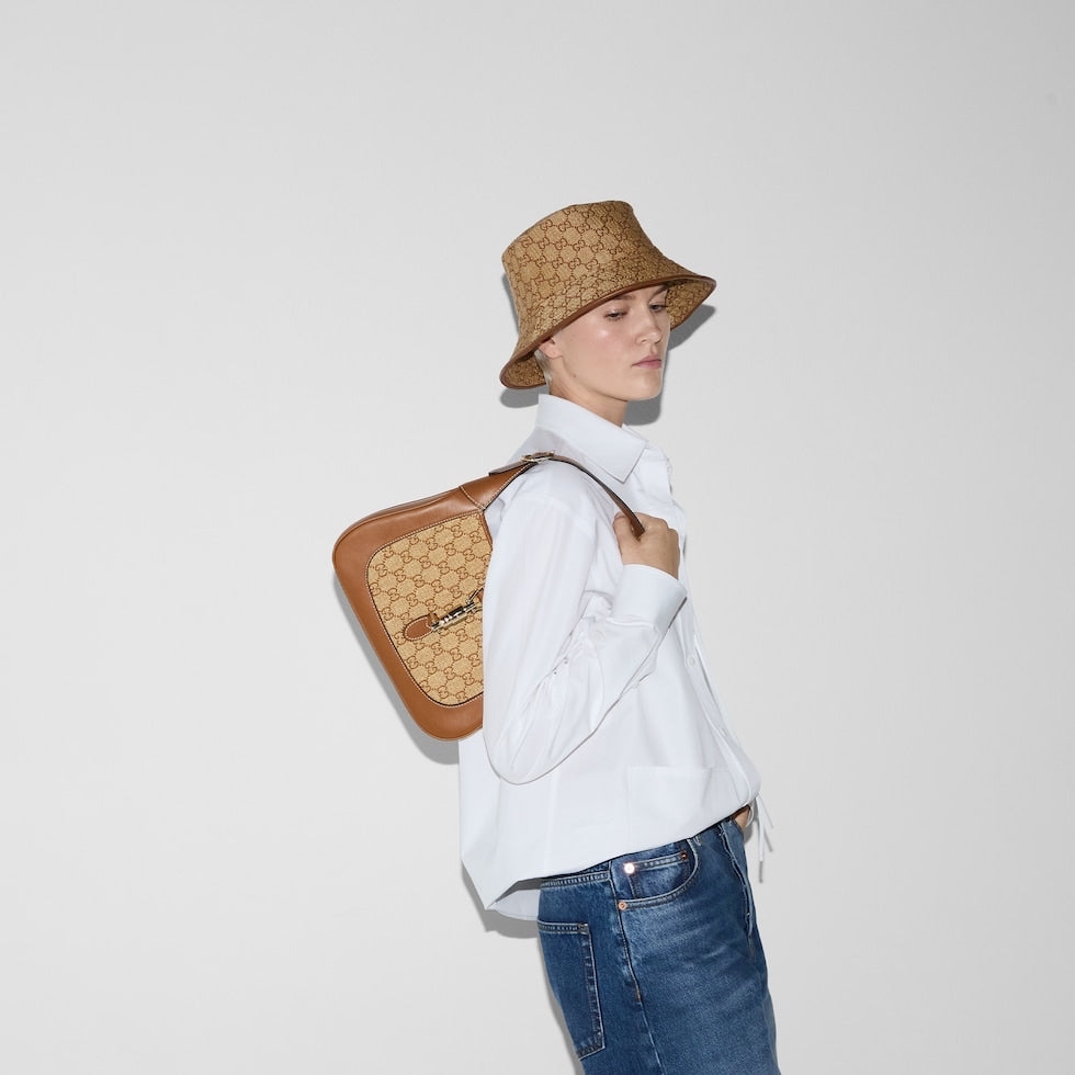 Person wearing a white shirt, blue jeans, and a brown bucket hat, holding a brown leather bag with a pattern.
