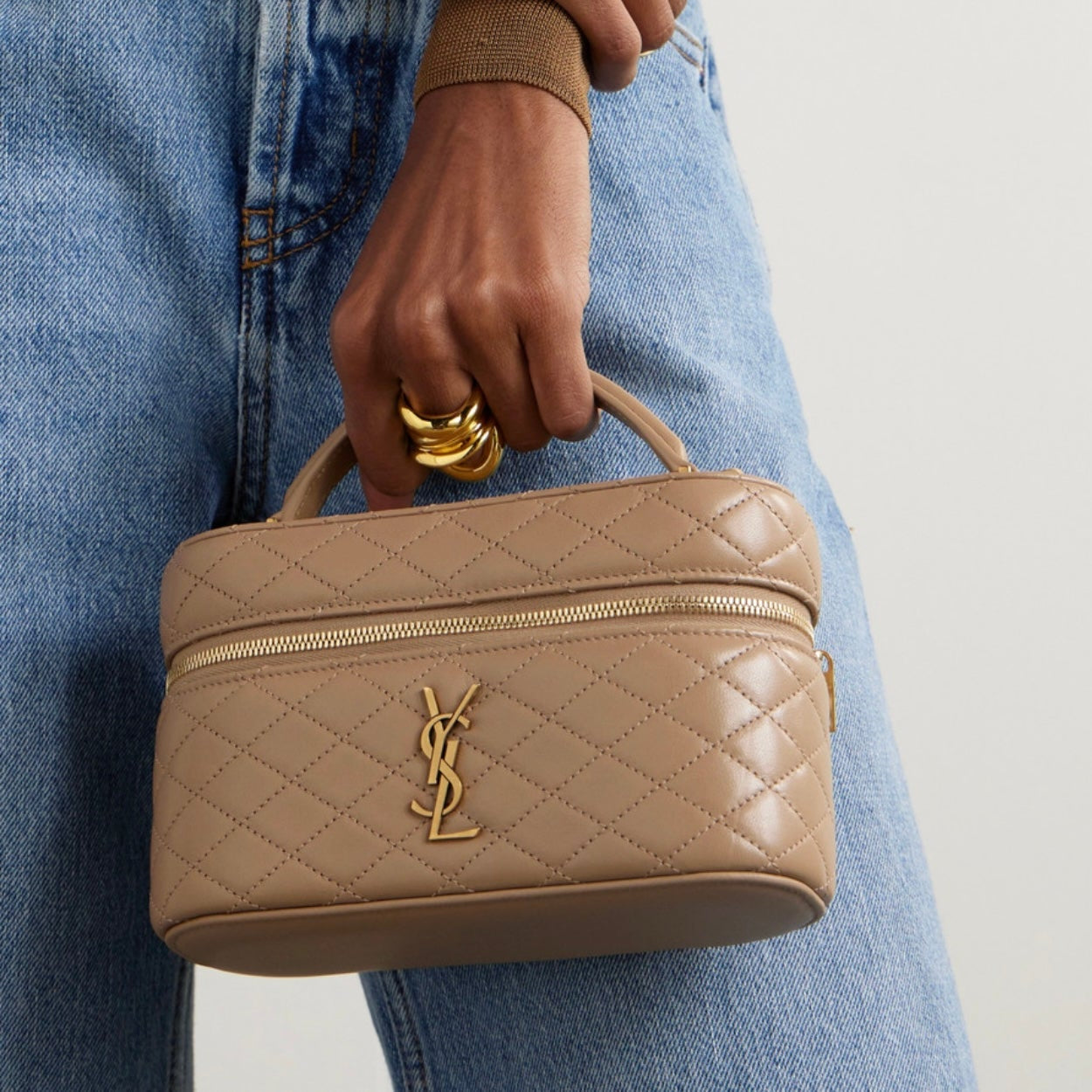 Person holding a beige quilted handbag with a brand logo, wearing blue jeans and a brown top.