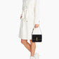 Person wearing a white trench coat holding a black handbag with a brand logo on a white background
