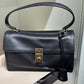 A black leather top-handle handbag with a gold-tone clasp and a hanging clochette, displayed on a store shelf next to other handbags.