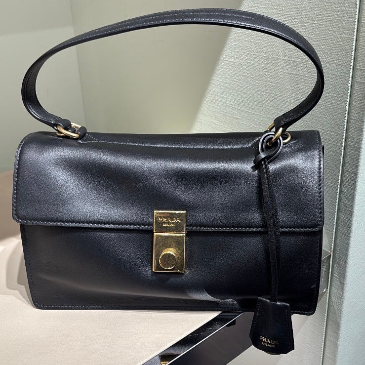 A black leather top-handle handbag with a gold-tone clasp and a hanging clochette, displayed on a store shelf next to other handbags.