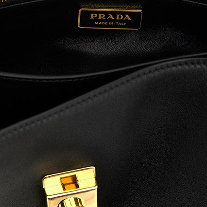 Close-up of a black leather Prada bag interior showing a gold-tone zipper, a gold Prada Made in Italy plaque, and a gold clasp on a black background.