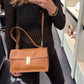 A woman takes a mirror selfie in a boutique while wearing a tan leather handbag with a gold clasp and shoulder strap. She is wearing a black textured knit sweater, black pants, and black flats.