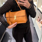 A woman takes a mirror selfie in a boutique while holding a tan leather shoulder bag. She is wearing a black knit sweater and black pants, with shelves of other handbags visible in the background.