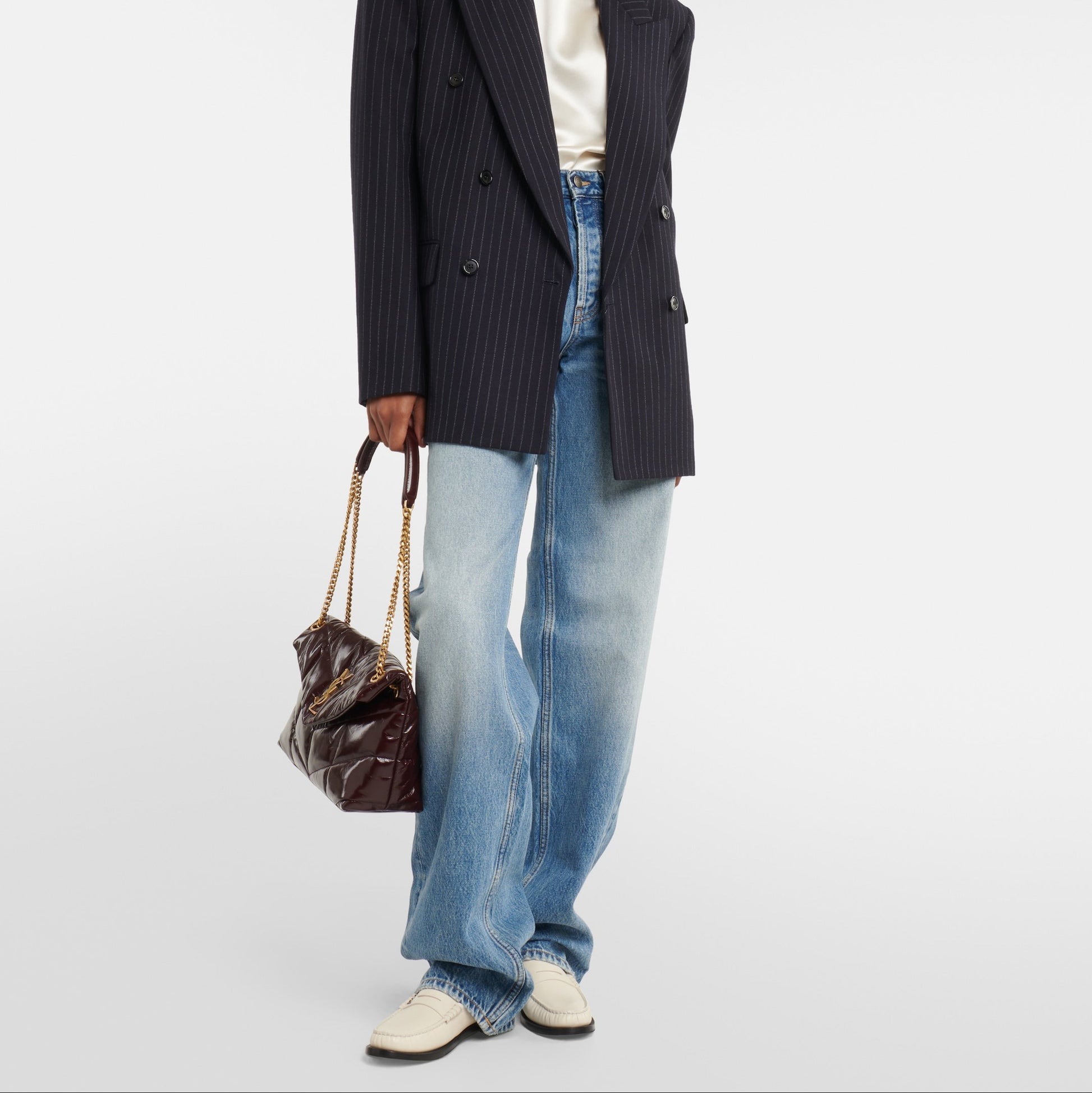 Person wearing a dark pinstripe blazer, white shirt, blue jeans, and holding a brown leather bag on a white background