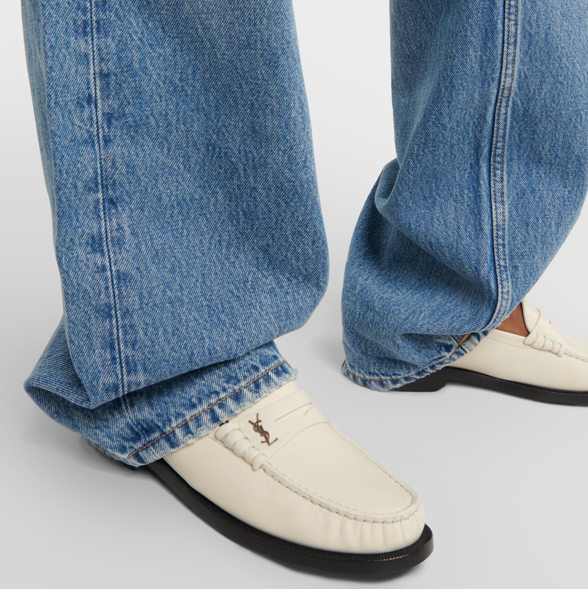 White loafers with a brand logo worn with blue jeans on a light gray background