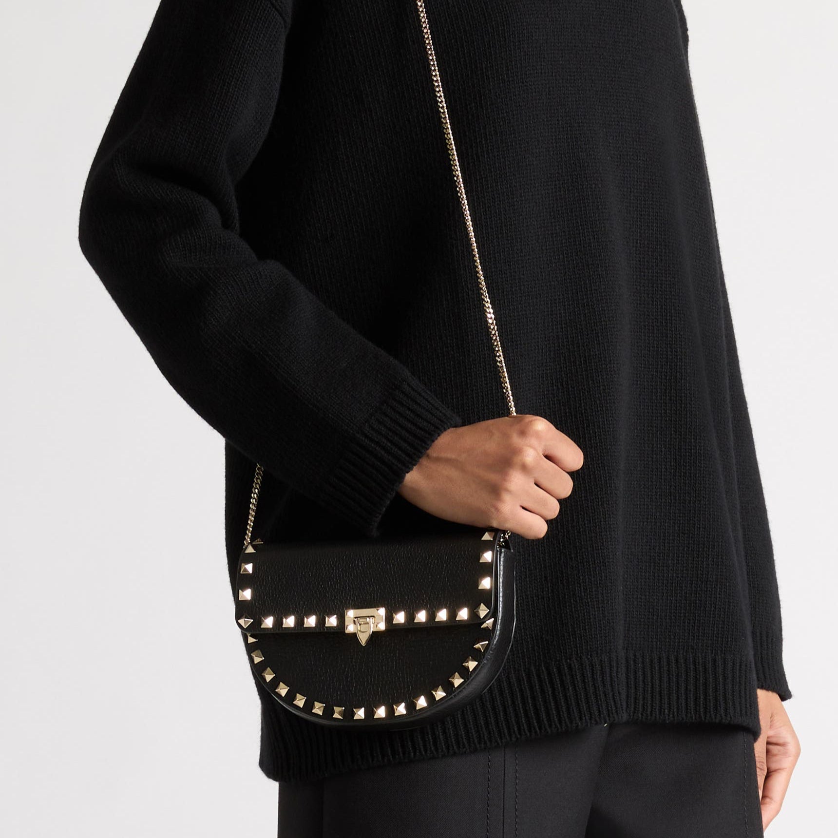 Person wearing a black sweater with a textured neckline, holding a black studded handbag against a white background.