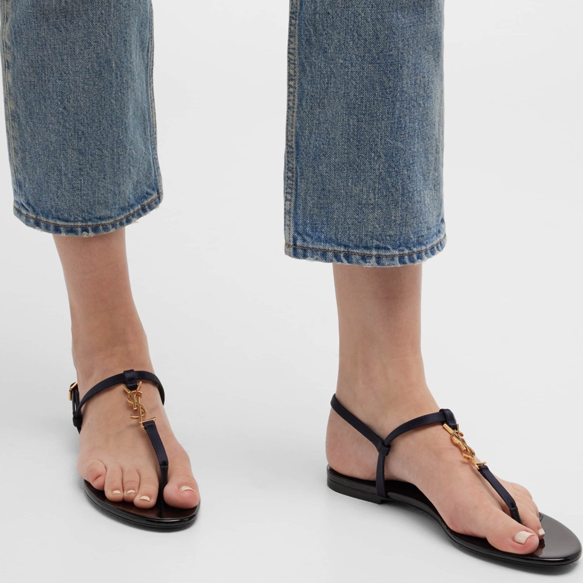 Black sandals with gold accents worn with rolled-up blue jeans on a white background