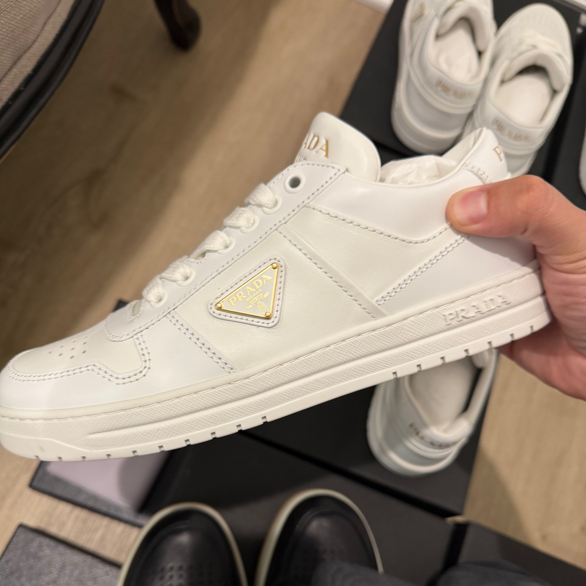 White sneaker with gold triangle logo held by a hand, with Prada branding visible.