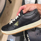 Black Prada sneaker with a yellow triangle logo held by a hand on a wooden floor.