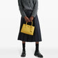 Person wearing a gray cable knit cardigan, white shirt, black skirt, and black shoes, holding a yellow handbag on a white background.