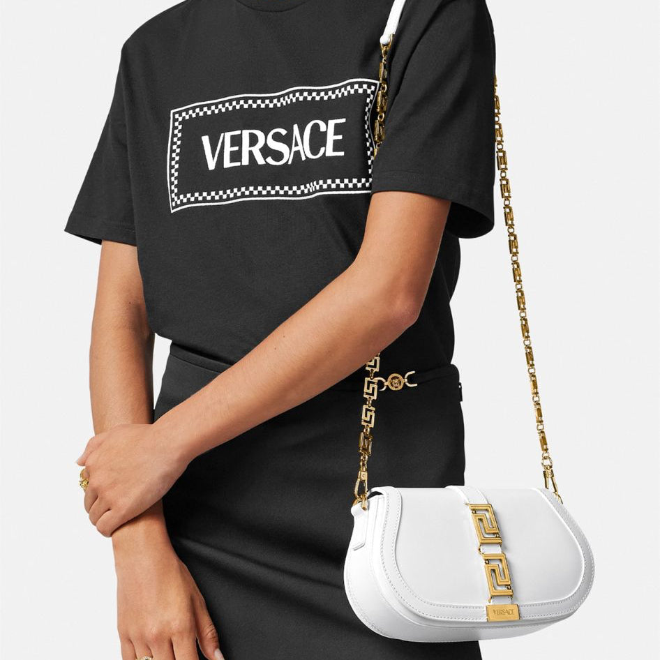 Person wearing a black Versace t-shirt with a white handbag featuring gold chain details.