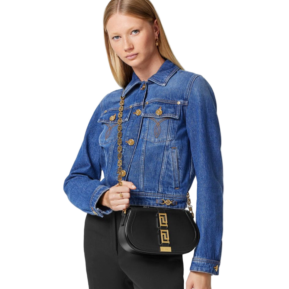 Woman wearing a blue denim jacket holding a black handbag with gold accents on a white background