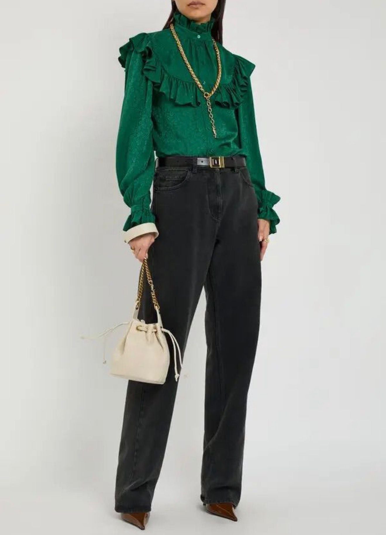 Person wearing a green blouse with ruffled details and black pants, holding a beige handbag on a white background.