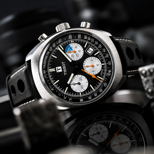 A Tissot Tixxom chronograph watch with a black dial, silver-tone stainless steel case, and a black perforated leather strap, shown in a dark, moody close-up with a reflection below.