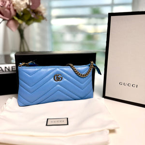 A small blue Gucci GG Marmont zipper pouch with a quilted design, placed on top of a white surface next to a black Gucci box and a floral arrangement.