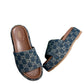 A pair of blue denim Gucci slide sandals with a platform sole and mid-heel, placed on a couch next to a green Gucci box.